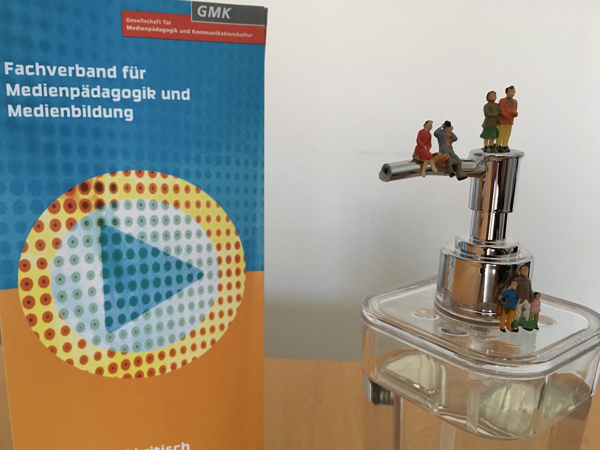 Foto eines Seifenspenders, auf welchem verschiedene Miniaturfiguren stehen und sitzen. Daneben steht ein Flyer der GMK.