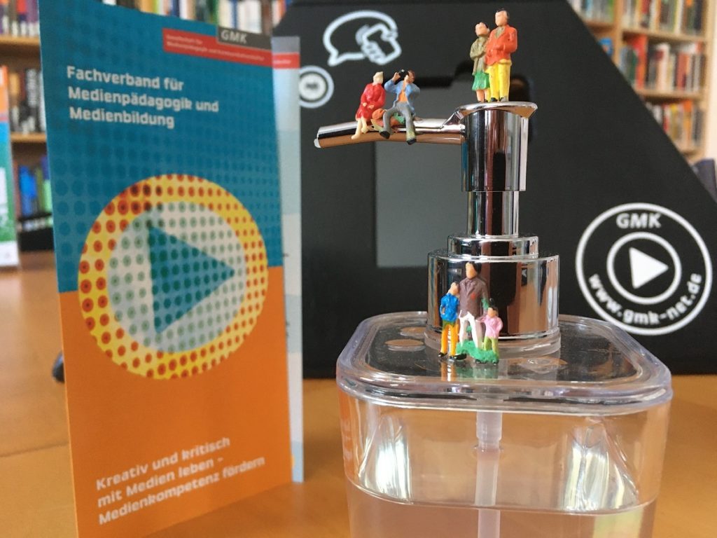 Foto eines Seifenspenders, auf welchem verschiedene Miniaturfiguren stehen und sitzen. Im Hintergrund steht ein Flyer der GMK.