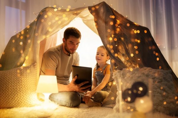 Foto einer Höhle aus Decken und Kissen, welche mit Lichterketten geschmückt ist. In ihr sitzen ein Mann und ein junges Mädchen im Schneidersitz und betrachten ein Tablet.