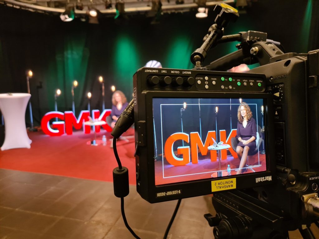 Foto eines Monitors, durch welchen man in das Studio des Forums Kommunikationskultur 2020 schaut. In dem Studio sitzt Anja Pielsticker vor den Initialen der GMK als dreidimensionale, weiß leuchtende Großbuchstaben mit roter Front