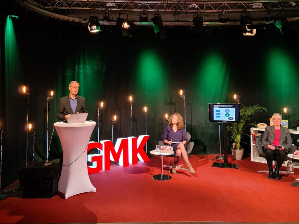 Foto des Studios des Forums Kommunikationskultur 2020. Im Studio sitzen Anja Pielsticker und Andrea Marten, welche Andreas Zick zugewandt sind, der an einem Stehtisch neben ihnen steht und spricht.