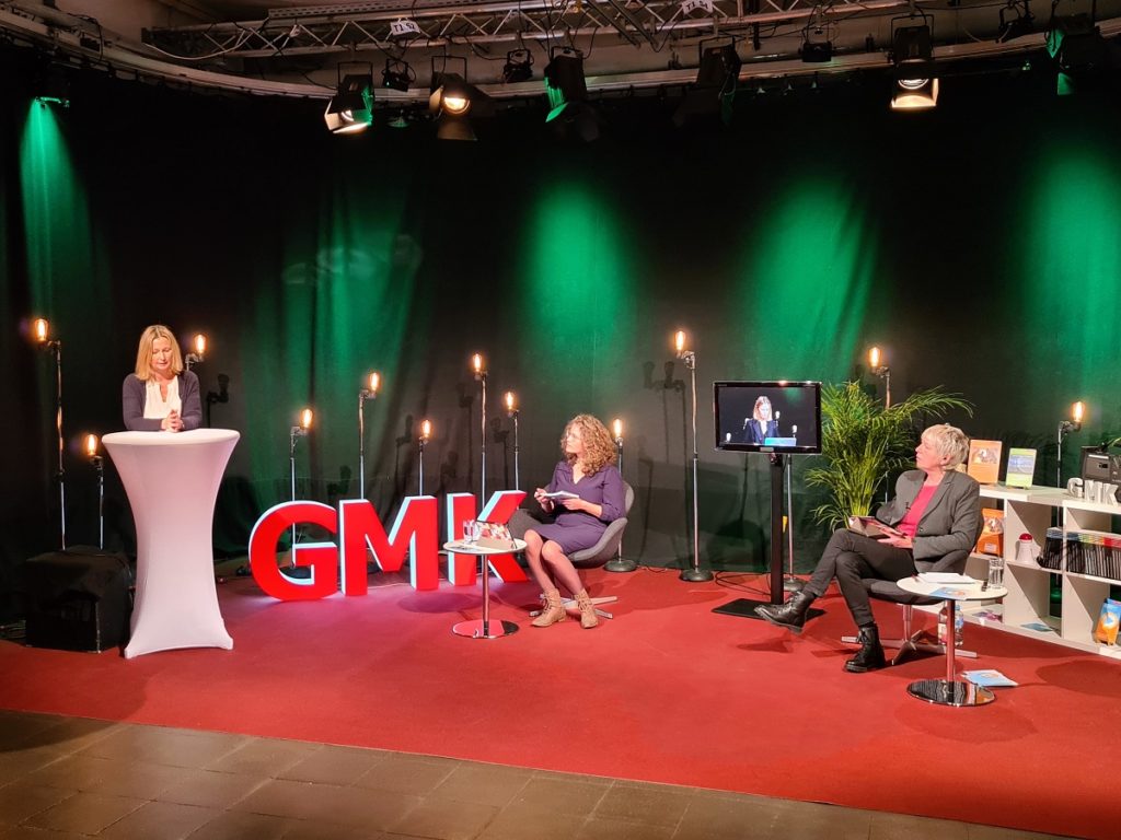 Foto des Studios des Forums Kommunikationskultur 2020. Im Studio sitzen Anja Pielsticker und Andrea Marten, welche Claudia Wegener zugewandt sind, die an einem Stehtisch neben ihnen steht und spricht.