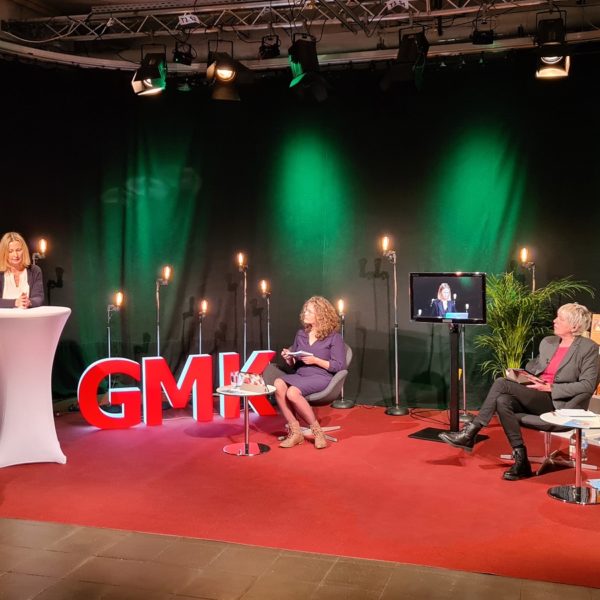 Foto des Studios des Forums Kommunikationskultur 2020. Im Studio sitzen Anja Pielsticker und Andrea Marten, welche Claudia Wegener zugewandt sind, die an einem Stehtisch neben ihnen steht und spricht.