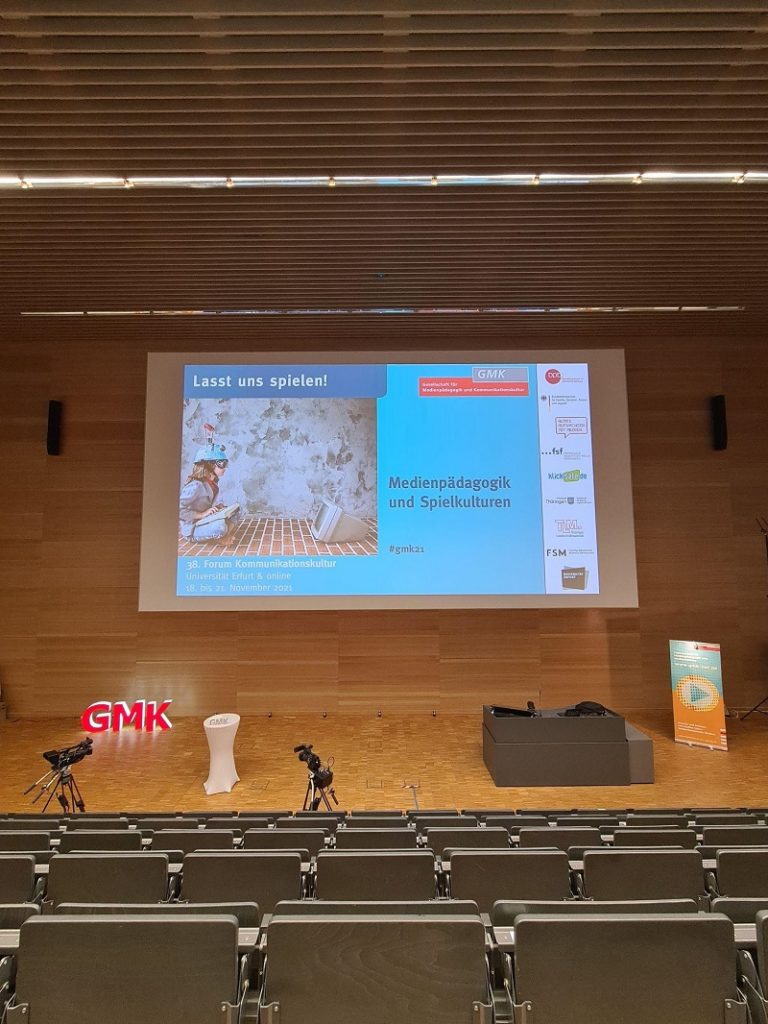 Foto des Hörsaals, in welchem das Forum Kommunikationskultur 2021 stattfand. Im Vordergrund sieht man ein paar Sitzreihen und im Hintergrund das Podium und die große Leinwand.