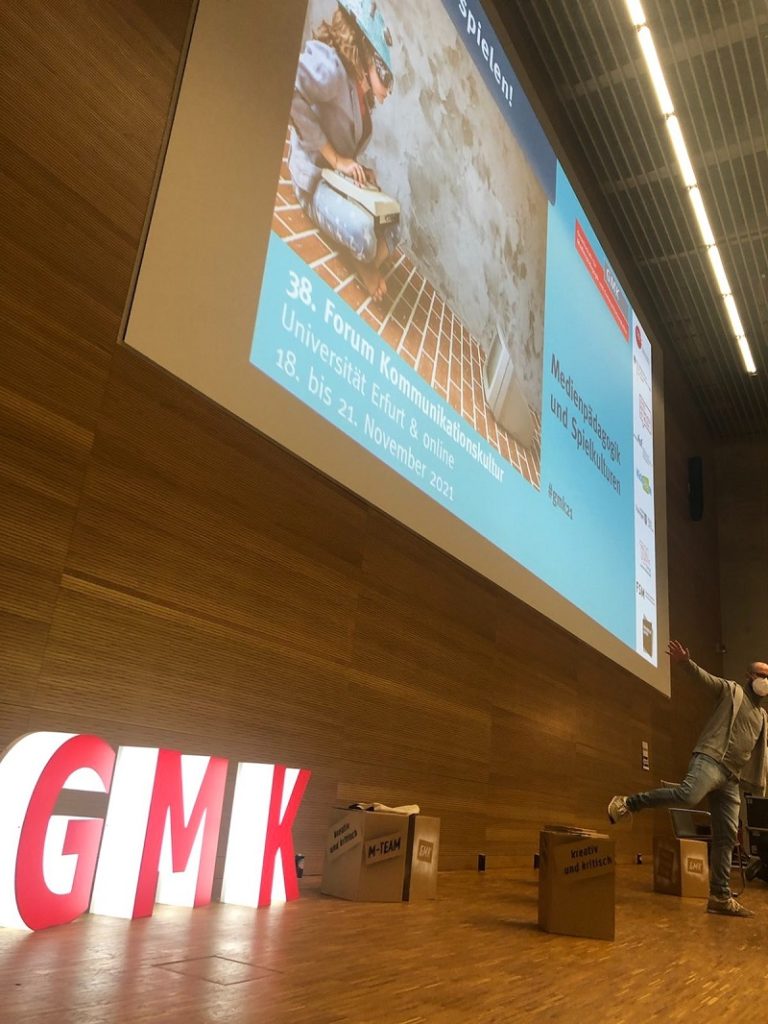 Von der Seite aufgenommenes Foto des Podiums des Forums Kommunikationskultur. Vor einer Holzvertäfelung stehen als dreidimensionale, weiß leuchtende Großbuchstaben mit roter Front die Initialen der GMK.