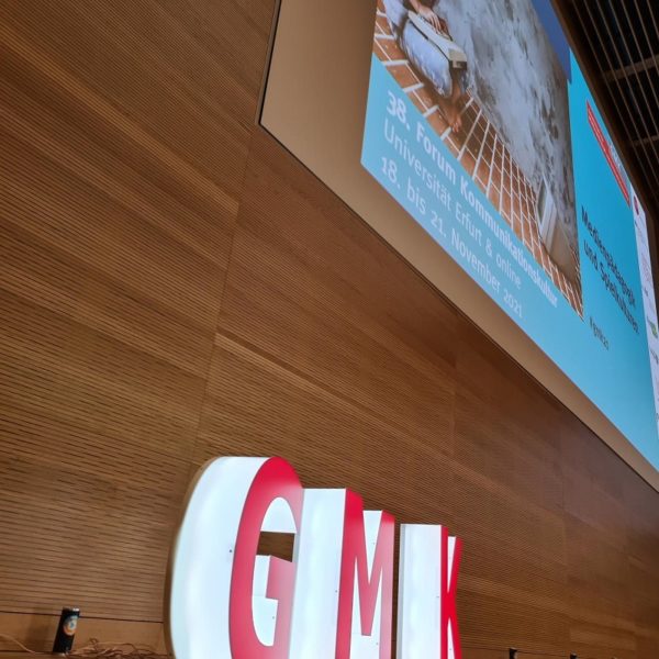 Von der Seite aufgenommenes Foto des Podiums des Forums Kommunikationskultur 2021. Vor einer Holzvertäfelung stehen als dreidimensionale, weiß leuchtende Großbuchstaben mit roter Front die Initialen der GMK.