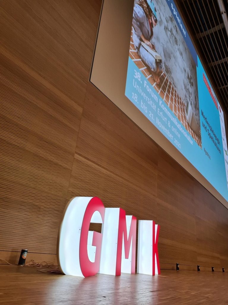 Von der Seite aufgenommenes Foto des Podiums des Forums Kommunikationskultur 2021. Vor einer Holzvertäfelung stehen als dreidimensionale, weiß leuchtende Großbuchstaben mit roter Front die Initialen der GMK.