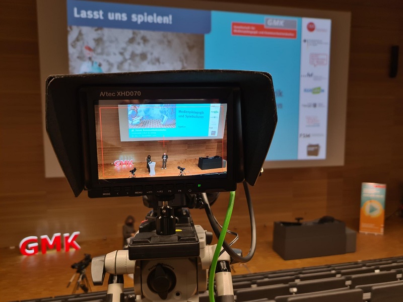 Foto eines Monitors, durch welchen man auf das Podium des Forums Kommunikationskultur 2021 schaut. Auf dem Podium steht das Moderationsteam Anja Pielsticker und Heiko Wolf.