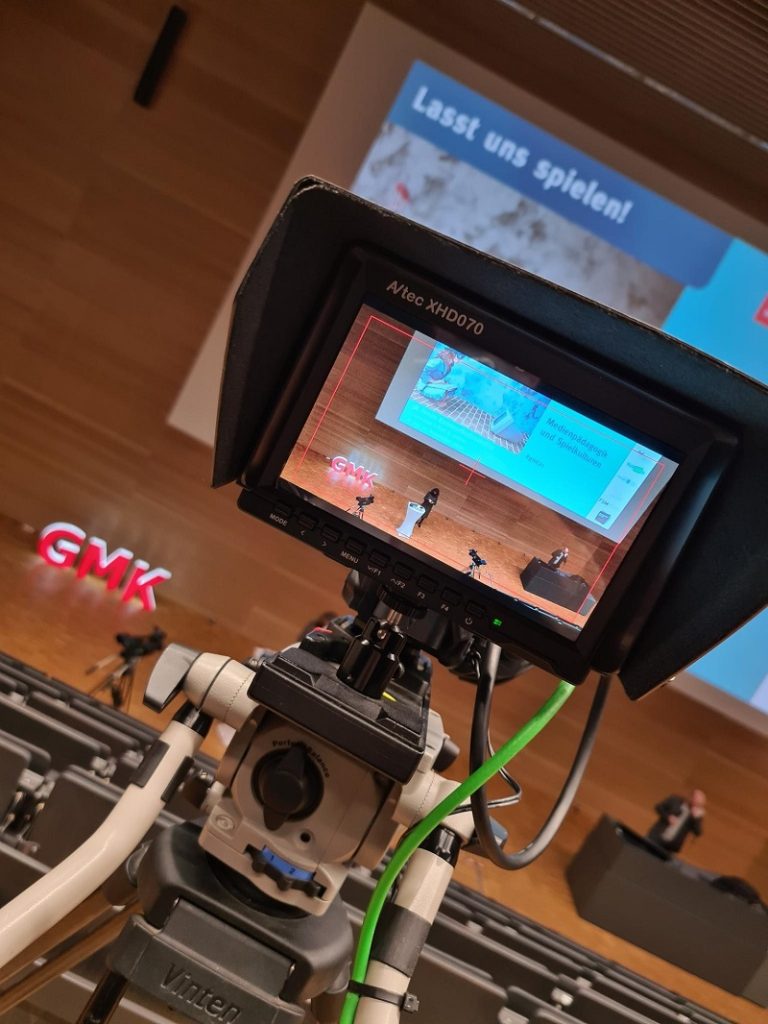 Foto eines Monitors, durch welchen man auf das Podium des Forums Kommunikationskultur 2021 schaut. Auf dem Podium steht das Moderationsteam Anja Pielsticker und Heiko Wolf.