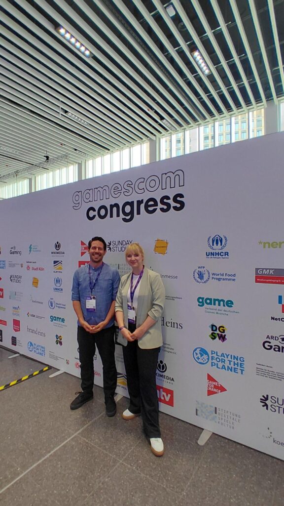 André Weßel und Linda Zeuner von der GMK stehen vor einer Wand mit dem Logo des gamescom congress und verschiedenen Partner*innenlogos