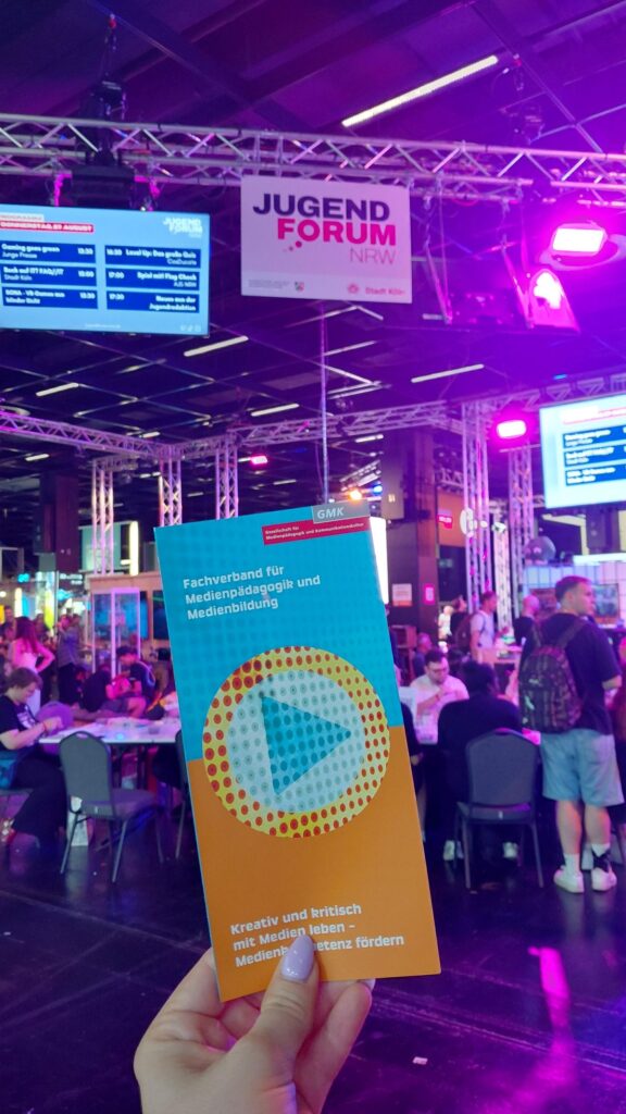 Person hält einen GMK-Flyer hoch. Im Hintergrund ist das Jugendforum NRW auf der Gamescom zu sehen